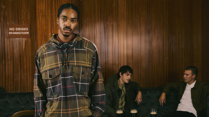Man in plaid shirt standing in a room with wooden paneling and other people sitting.